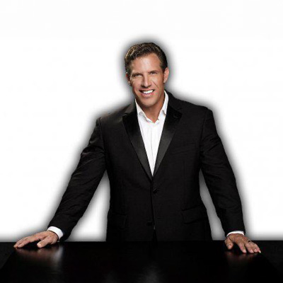 The image features a man dressed in formal attire, standing behind a podium with his hands resting on its surface.