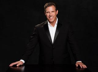 The image features a man standing at a podium, dressed in formal attire with a tuxedo and bow tie, smiling and looking directly at the camera.