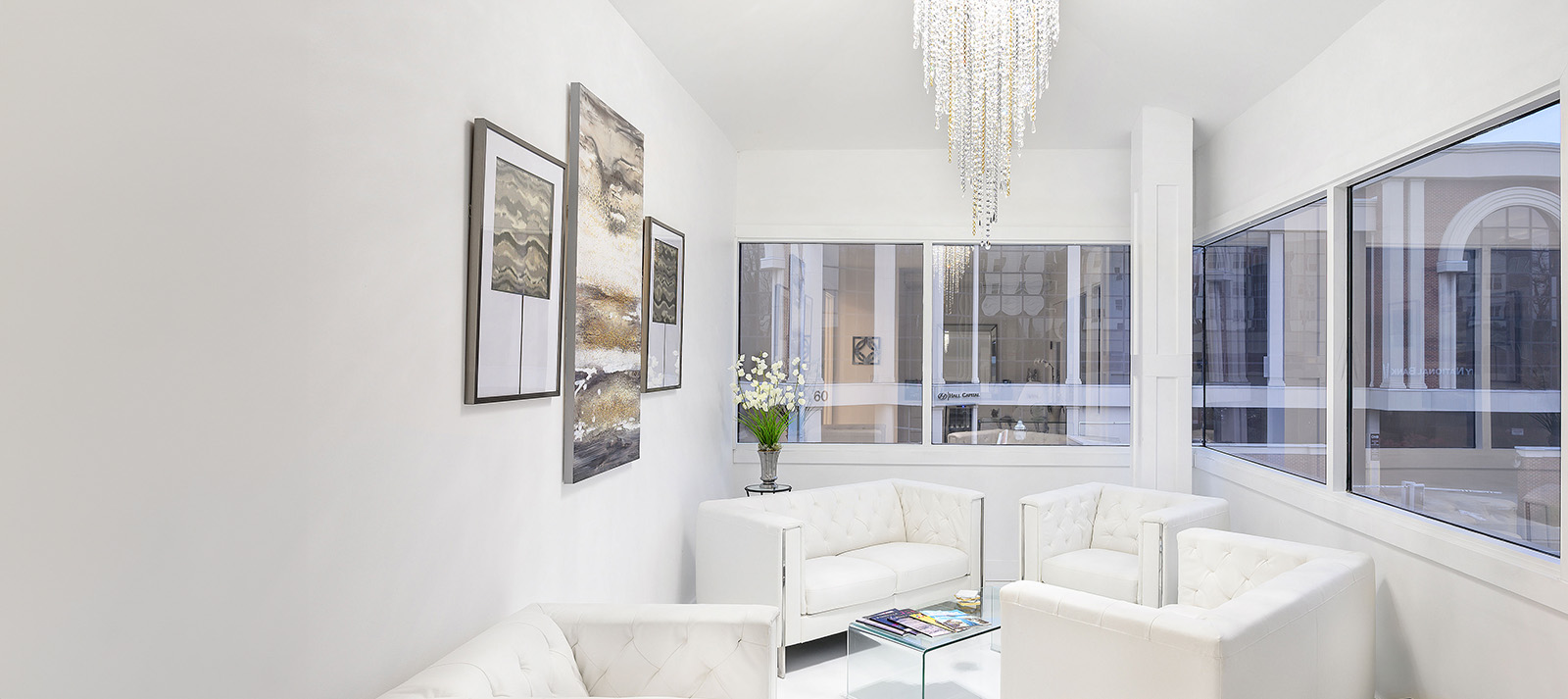 A modern living room with white walls, large windows, a chandelier, white furniture, and a view of a city street through the windows.
