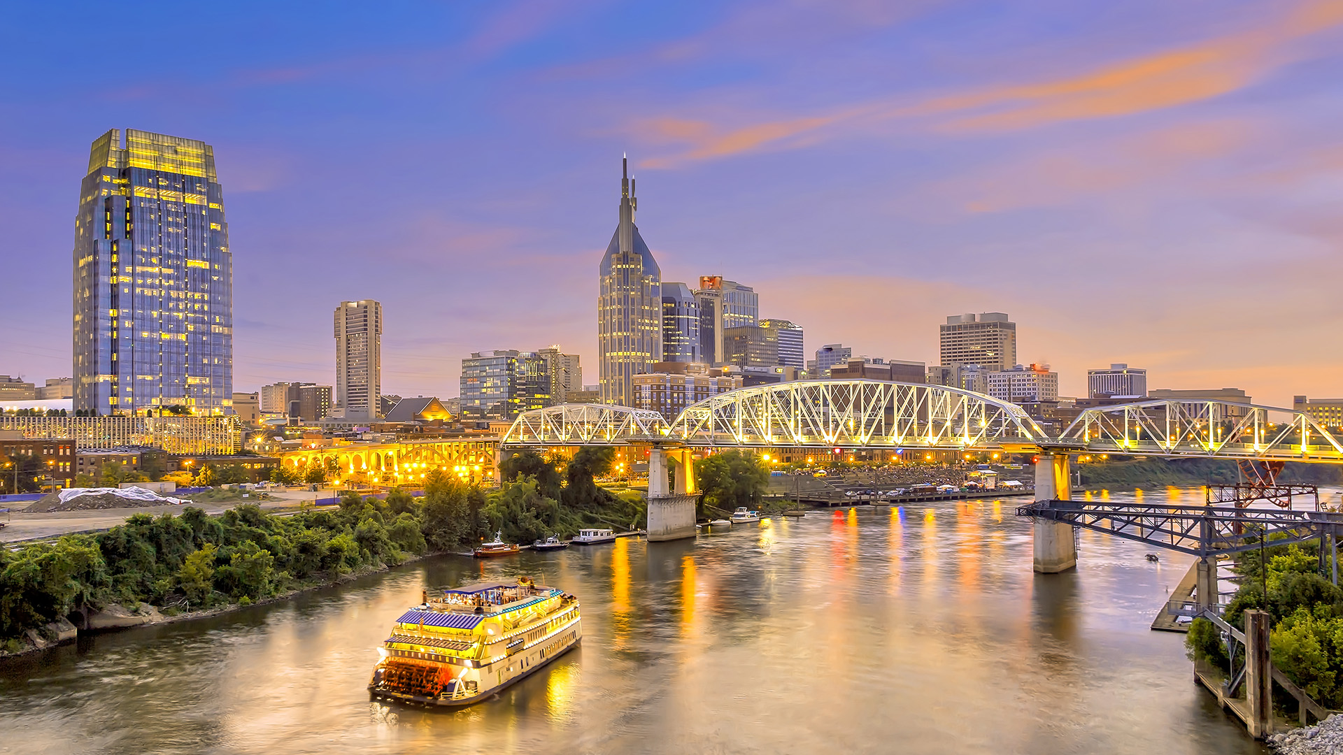 The image depicts a city skyline with a prominent river bridge at night, with a boat on the waterway and a large building illuminated in the background.