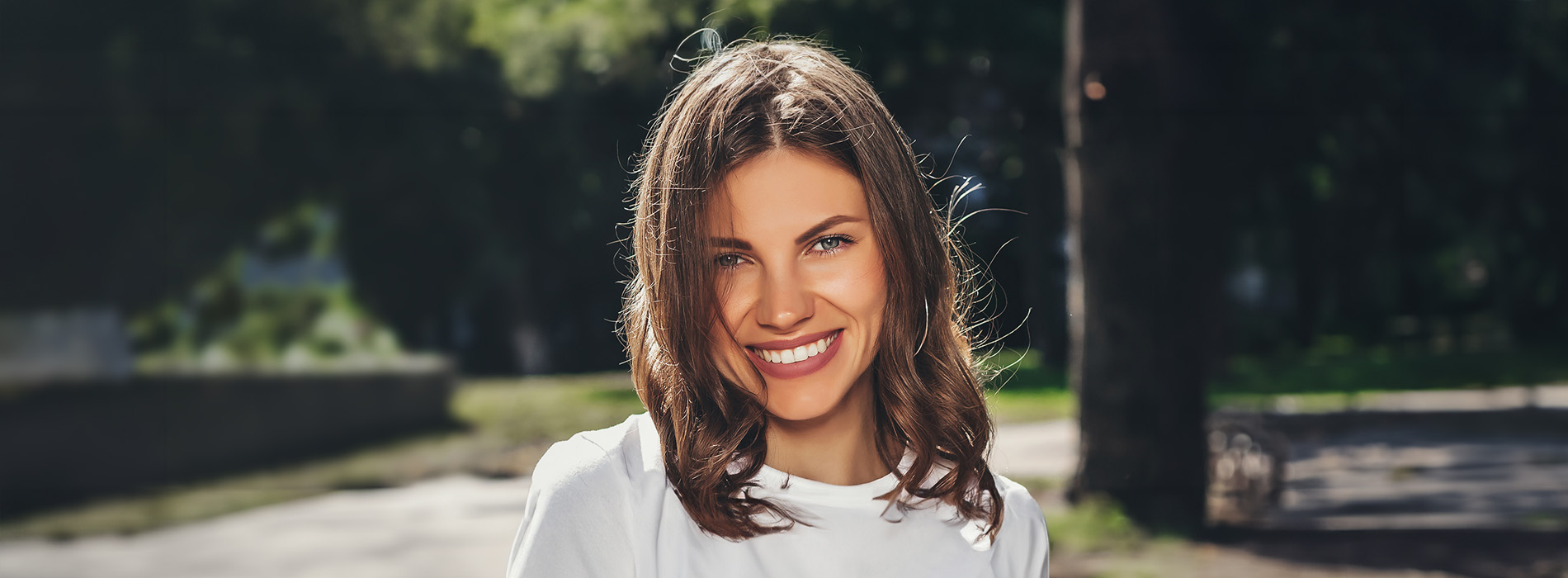 A young woman with long hair smiling at the camera, wearing a white top against an outdoor background.