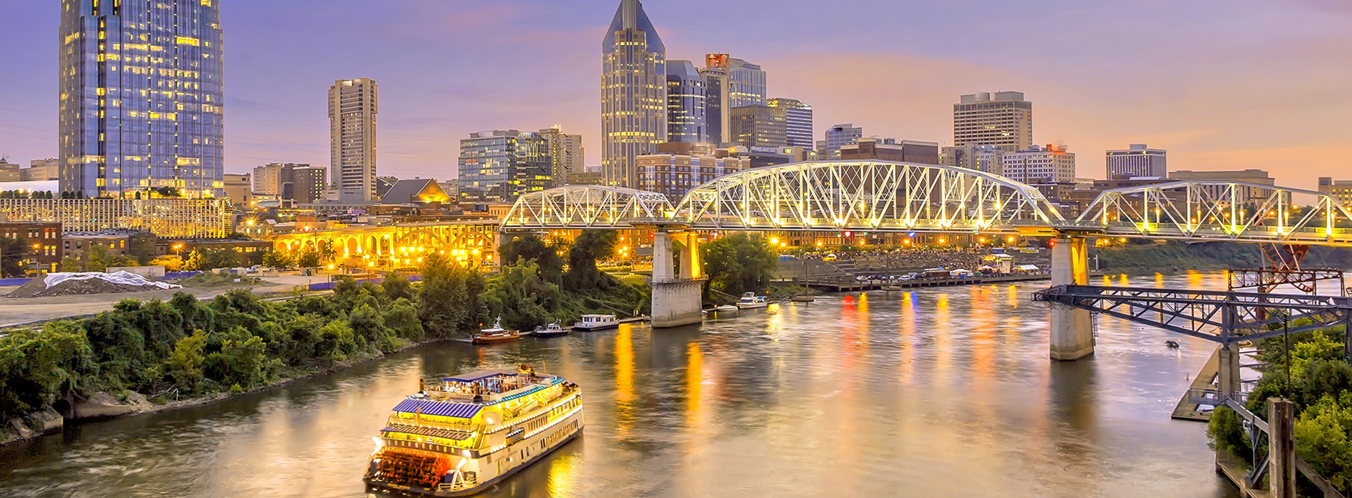The image depicts a city skyline at dusk with illuminated buildings, a river with boats, and a bridge spanning across the water, all under a colorful sky.