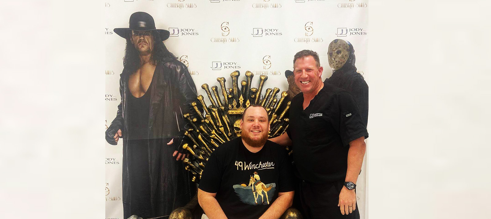 The image shows two men posing with a life-size cardboard cutout of a person dressed as a superhero, standing in front of a backdrop that includes a character from a TV show and other promotional items.