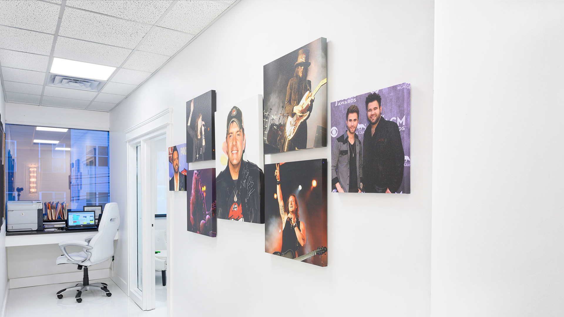 The image shows a wall with multiple framed photographs displayed, likely within an office setting, with a person seated at a desk visible in the background.