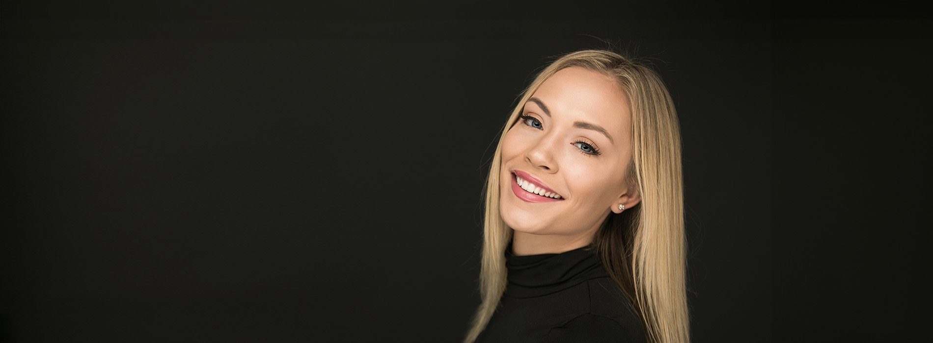 The image shows a woman with blonde hair smiling towards the camera against a dark background.