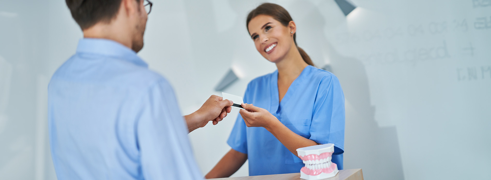 A man and woman in a professional setting, with the man handing a card to the woman who appears to be a receptionist, both standing behind a counter.