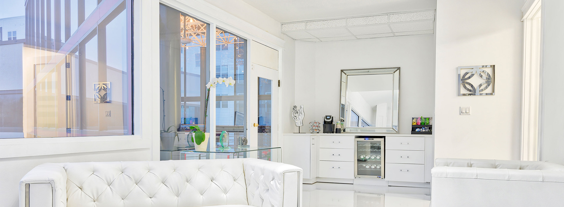 The image displays a modern interior space with a large open floor plan, featuring a living room area with a white sofa, a sleek coffee table, and a contemporary design aesthetic, leading into a dining area with a glass-topped table and chairs, which transitions to a kitchen area equipped with stainless steel appliances and a clean countertop. The space is well-lit and appears to be part of a showroom or model home.
