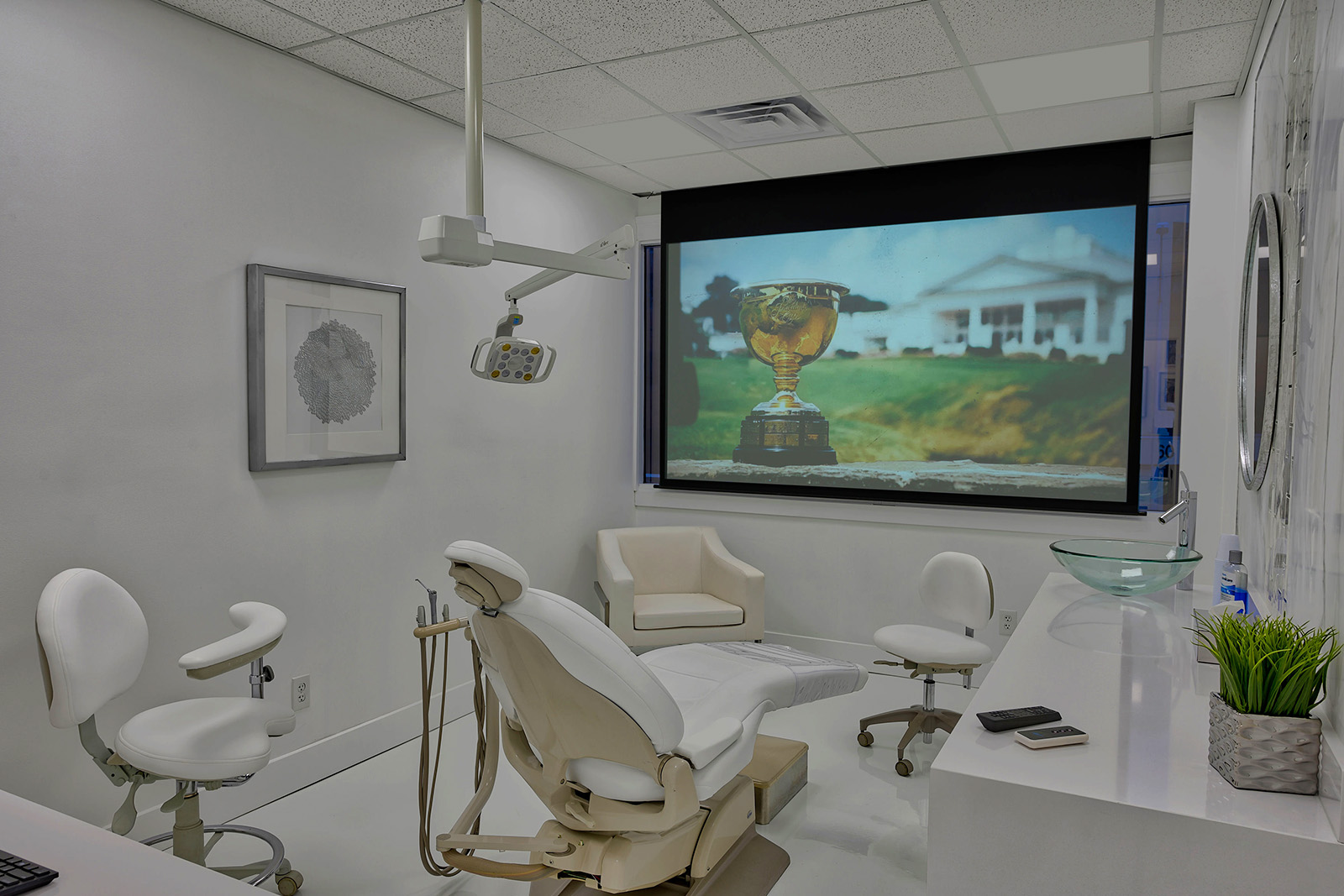 The image shows an interior view of a dental office with various pieces of equipment and a large screen displaying a golf trophy on a pedestal.