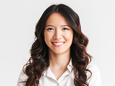 A smiling woman with dark hair and bangs, wearing a white shirt and standing against a neutral background.