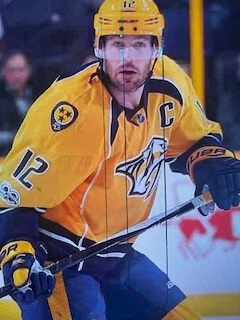 Hockey player in yellow jersey with  nashville  written across chest, holding hockey stick on ice, smiling towards camera.