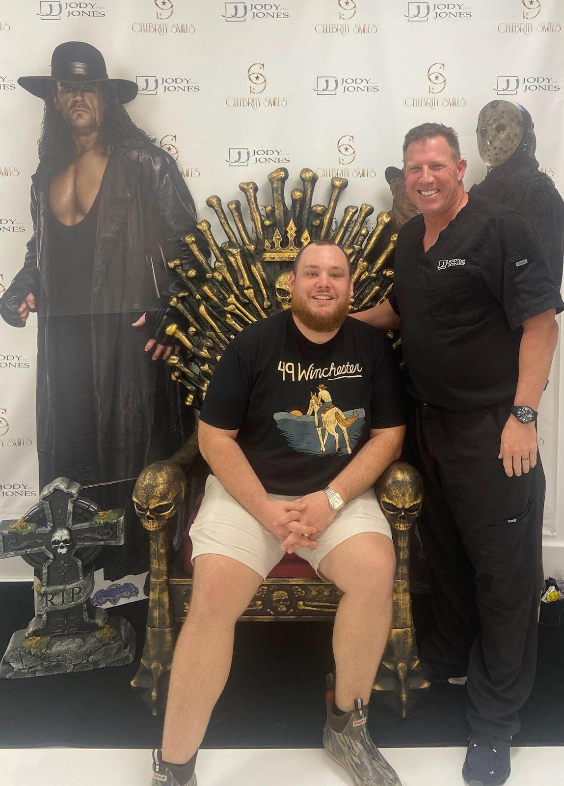A man and another person posing next to a life-sized replica of the Iron Throne from Game of Thrones, with a backdrop featuring cutouts of characters from the show.