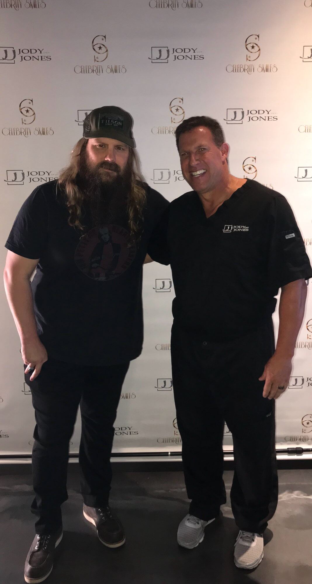 The image features two men posing together; one is wearing a black t-shirt and sunglasses, while the other is dressed in scrubs and a white coat, standing next to a promotional sign.