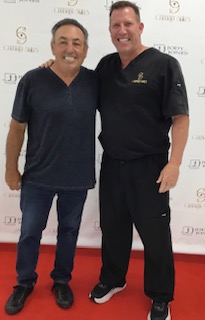 In the image, there are two men standing side by side on a red carpet. The man on the left appears to be wearing casual clothing while the man on the right is dressed in professional attire, suggesting they may be at a formal event or gathering.