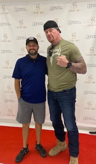 This image features a man with a beard wearing a black tank top standing next to another man who is wearing a camouflage shirt and a baseball cap, posing for a photo together.