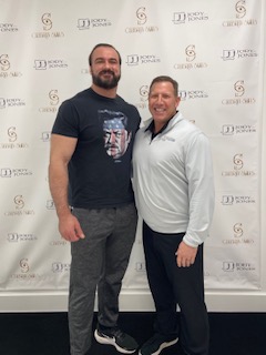 The image features two men standing side by side against a backdrop with text  the man on the left is wearing a black t-shirt with a graphic and has short hair, while the man on the right is dressed in a white shirt with a logo and dark pants, and he has graying hair.