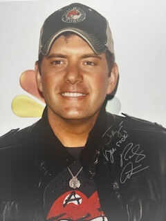 The image shows a man posing with a smile, wearing a baseball cap, a t-shirt, and a necklace, against a backdrop that includes a logo and text.