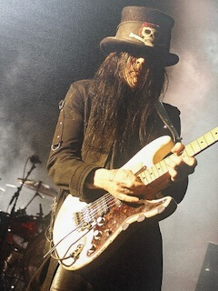 The image shows a person playing an electric guitar on stage, dressed in a distinctive black and white costume with a top hat and sunglasses, resembling a character that could be associated with heavy metal music culture.