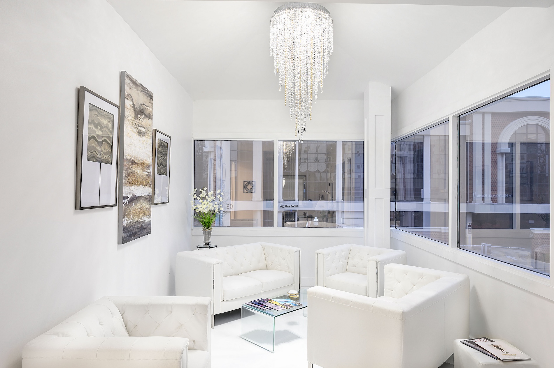 The image shows a modern living room interior with white walls, a large chandelier, contemporary furniture including a sofa and chairs, and a framed artwork on the wall.
