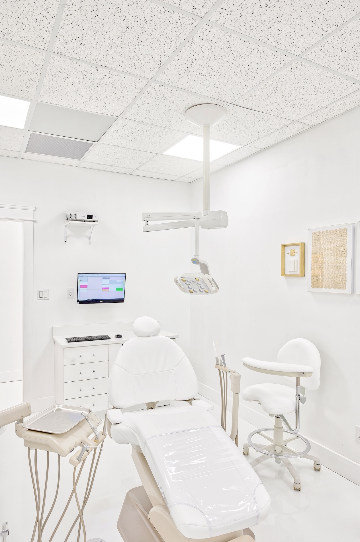 The image shows a well-equipped dental office with a clean and modern appearance, featuring a dental chair, examination table, adjustable lighting, and various medical equipment.