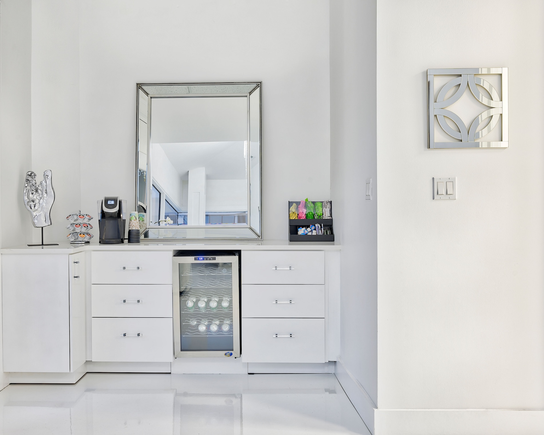 The image shows a well-lit modern bathroom interior with white walls, a large mirror above a sink, a white cabinet below the sink, a stainless steel refrigerator, a white countertop, and a silver metal towel rack on the wall.