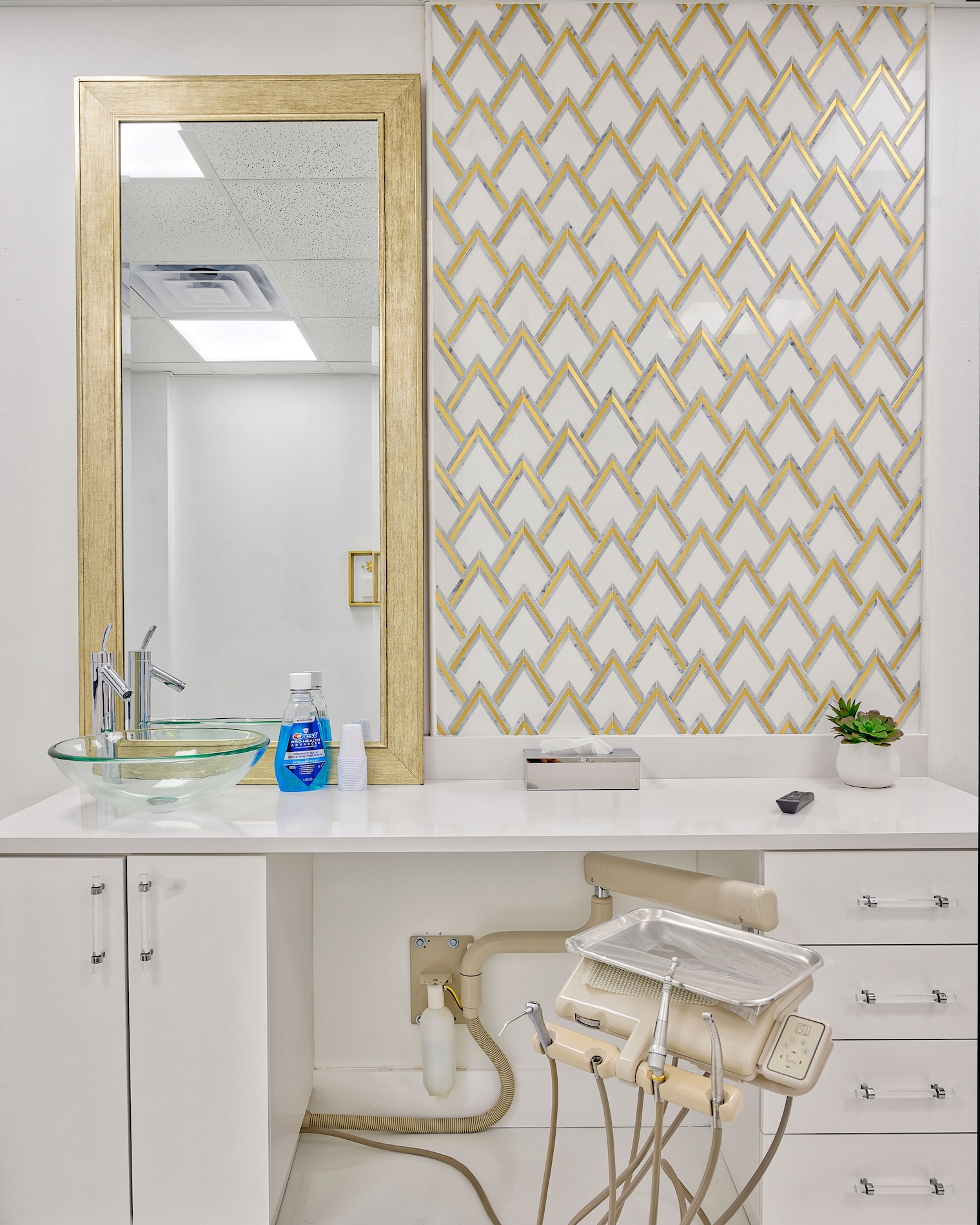 The image shows a bathroom vanity area with a gold geometric patterned wall, a white sink, a mirror above the sink, a dental chair on the right side, and various items on the countertop such as a bottle of mouthwash and other dental care products.