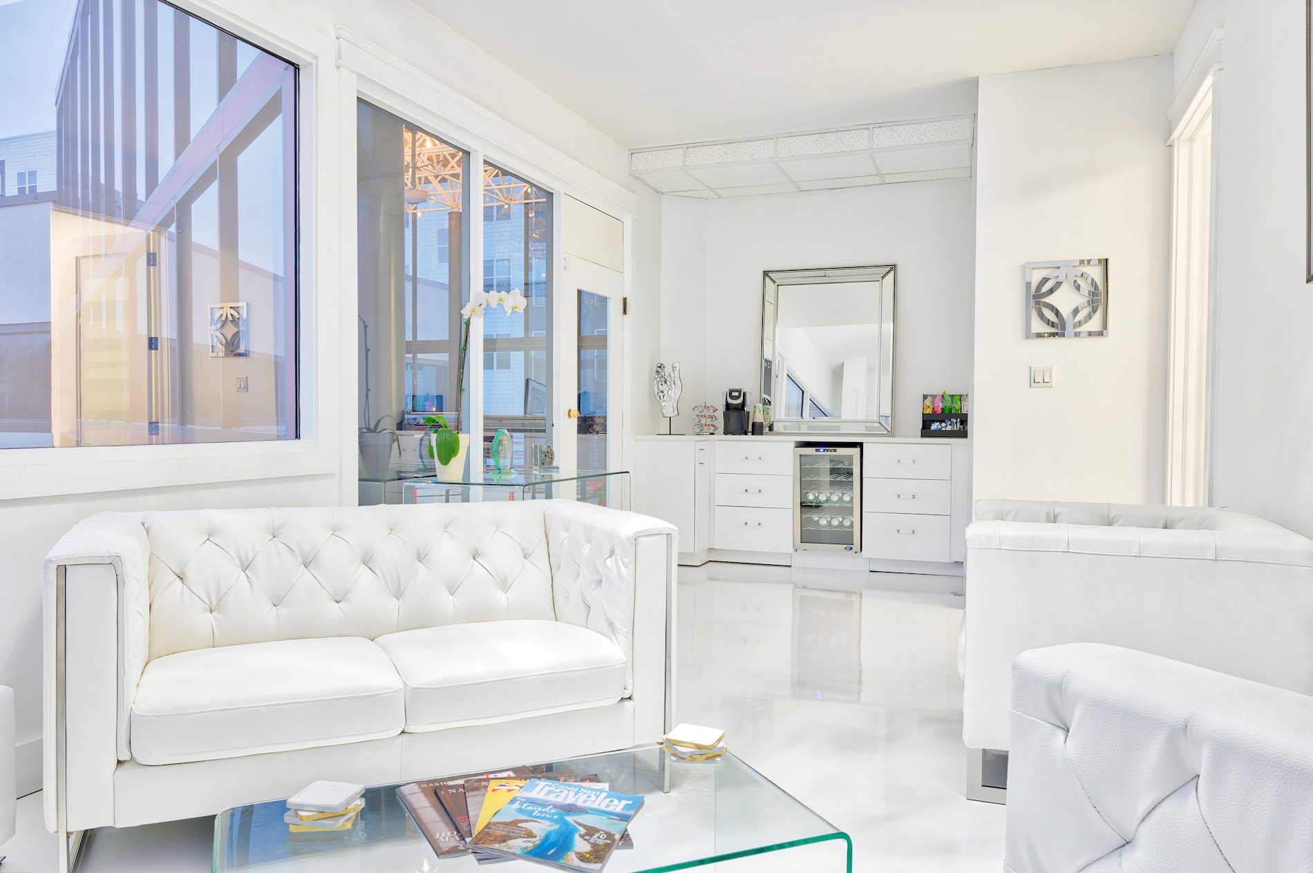 The image shows a modern interior space with a white sofa, a sleek kitchenette, a dining table, and large windows allowing natural light to enter.