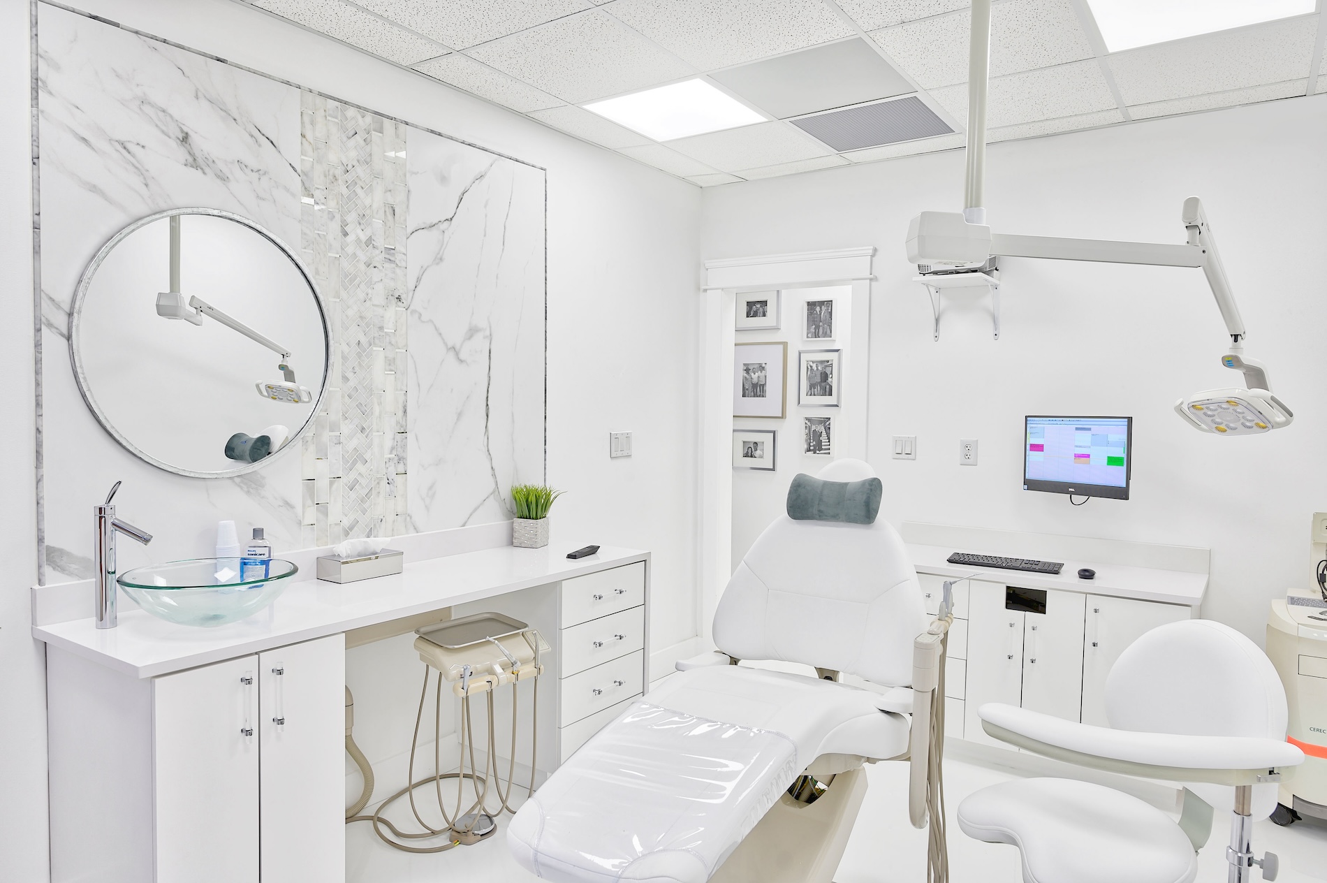The image shows an interior view of a dental office with modern furnishings and equipment, featuring a reception desk, dental chair, mirror, sink, and various dental tools.