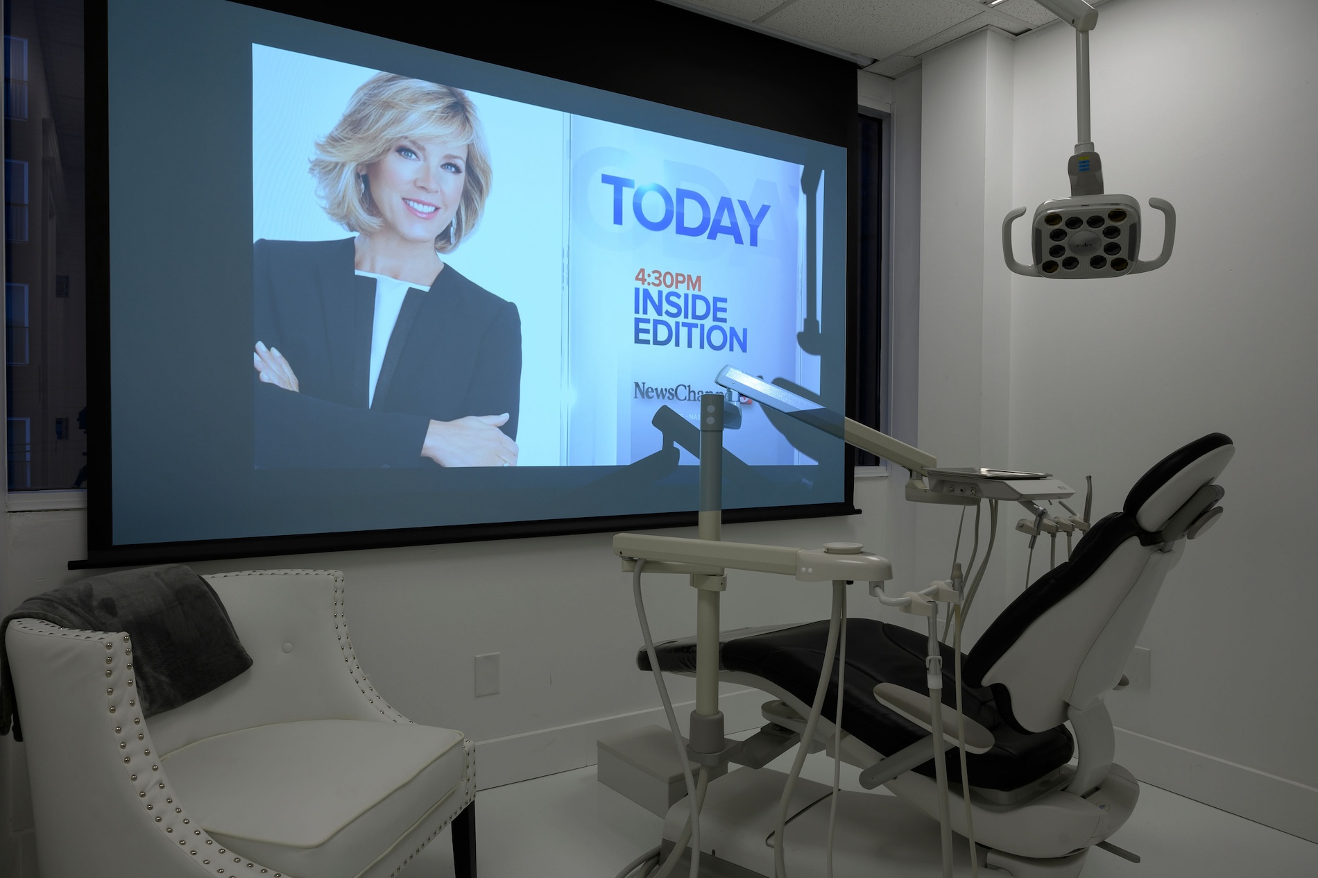 The image shows an interior space that appears to be a dental office with various pieces of equipment visible, including a dental chair and a large screen displaying a news program featuring a woman presenter.