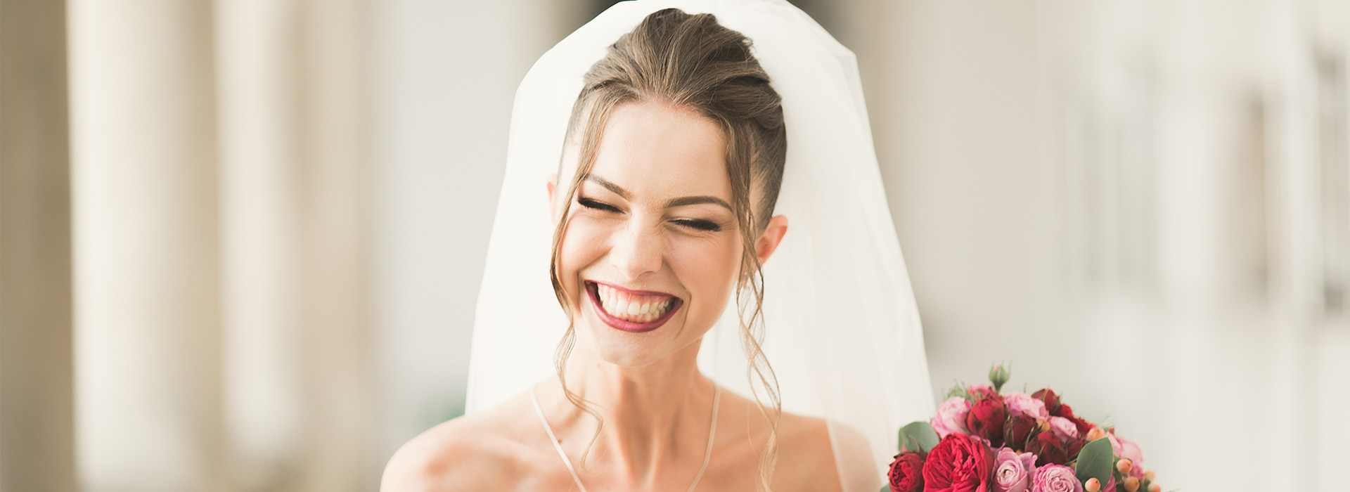 A bride with a radiant smile, wearing a white veil and necklace, stands next to a bouquet of red flowers at a wedding venue.
