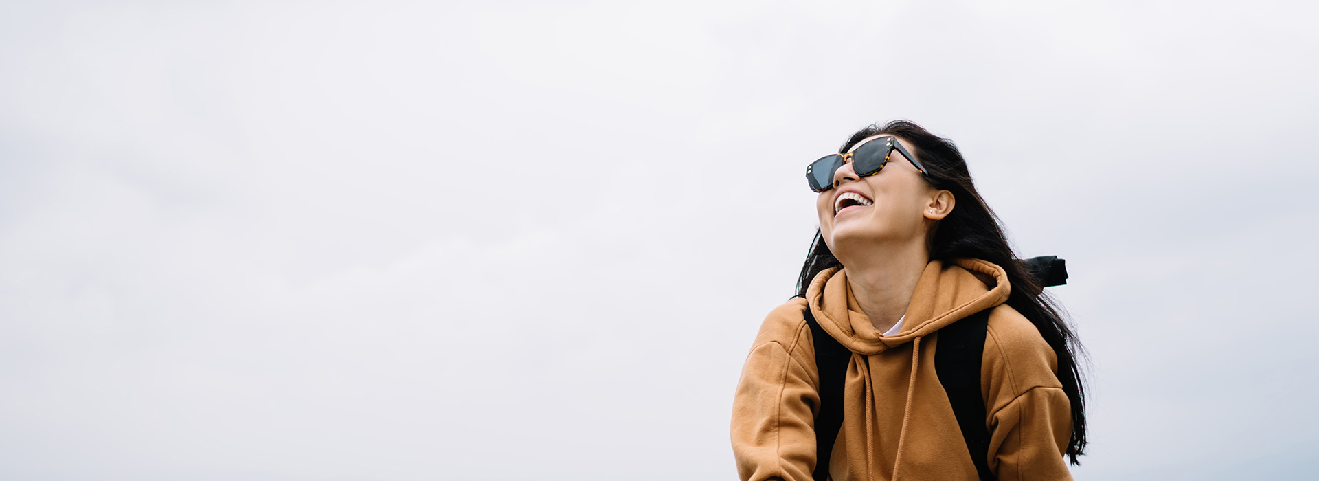 A person standing outdoors with sunglasses, smiling, wearing a backpack, and looking towards the camera.