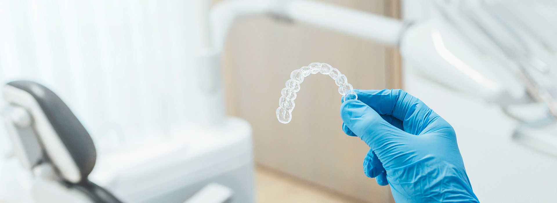 The image shows a person s hand holding a transparent dental impression material over a dental chair with equipment, suggesting a dental care setting.