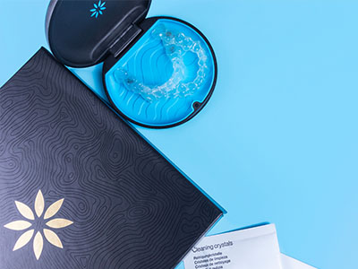 The image shows a closed blue box with a white interior, containing a transparent object that appears to be a dental impression tray, next to an open package of dental floss and a toothbrush on a blue surface.