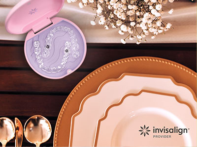 The image shows a collection of tableware on a wooden surface, including plates, bowls, and a lid with a design resembling a seashell, accompanied by a boxed product that appears to be dental care-related, all set against a blurred background featuring a floral arrangement and a pink and white color scheme.