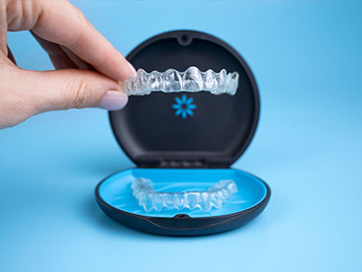 The image shows a hand holding an open dental retainer case containing clear aligners, with a blue lid partially open and a person s reflection visible in the transparent plastic tray.