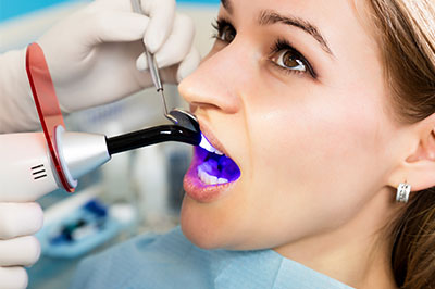 The image shows a person sitting in a dental chair with a device attached to their mouth, likely for a dental procedure, while wearing a surgical mask and being attended to by a professional who appears to be using a machine with a blue light for treatment.