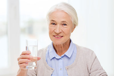 The image features an elderly woman holding a glass of water with her right hand, smiling gently at the camera.
