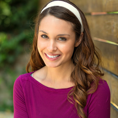 A young woman with long hair, wearing a purple top and a headband, smiles at the camera against a wooden fence background.