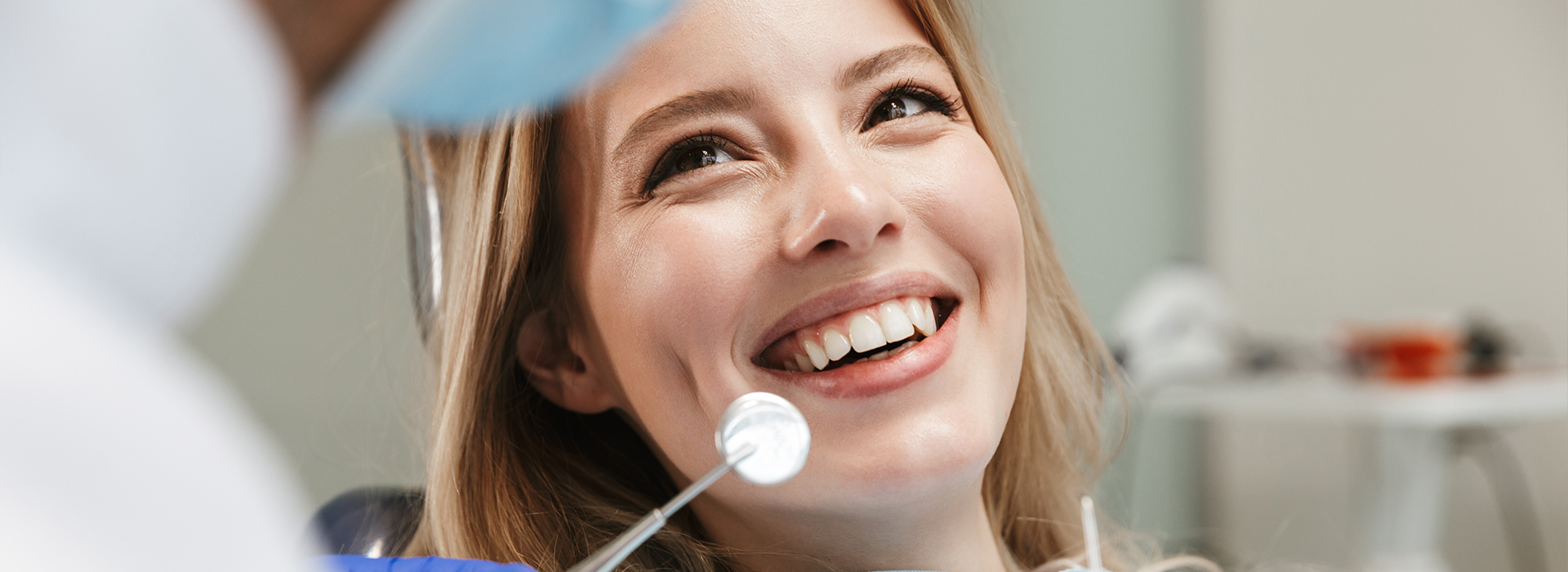 A smiling woman with blonde hair sitting in front of a dental professional, both wearing face masks, with dental tools visible in the background.
