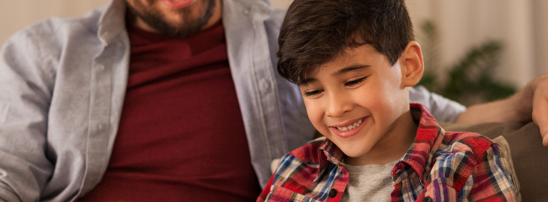 A man and a young boy are seated on a couch, smiling at each other while looking directly into the camera.
