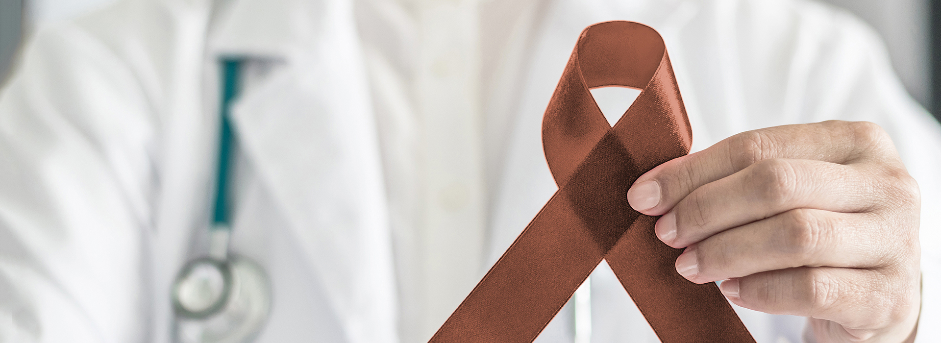 The image shows a person holding a red ribbon with a white cross symbolizing breast cancer awareness, while wearing a white lab coat and stethoscope, suggesting a medical professional in the context of healthcare.