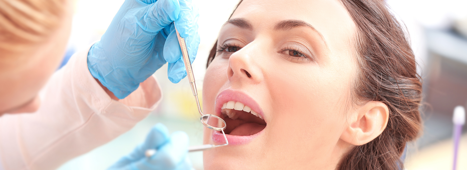This is an image featuring a woman receiving dental care, with a dentist performing a procedure on her teeth while she is seated in a dental chair, wearing a blue surgical gown and a face mask.