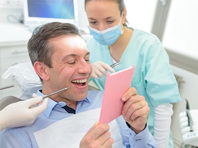 The image shows a man sitting in a dental chair with a pink cardboard sign in front of him, smiling at the camera, while a female dentist stands behind him, holding a toothbrush and looking at the card.