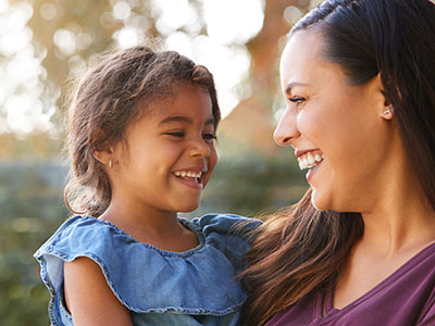 A woman and a young girl are smiling at each other, with the adult holding the child close, both outdoors during daylight.