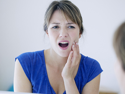 The image shows a woman with her mouth open, looking concerned or possibly in pain, with a hand placed on her chin. She appears to be indoors, with a mirror reflecting her face.