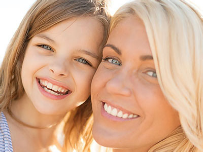 The image shows a woman and a young girl smiling at the camera, with the woman appearing to be middle-aged and the child having blonde hair. They are outdoors, possibly on a beach, given the presence of sand in the background.