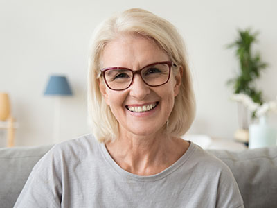A smiling woman with short blonde hair, wearing glasses and a gray top, seated indoors.