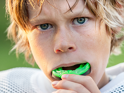 The image shows a young boy with blonde hair, wearing a football uniform with his mouth open, holding a green object up to his face that appears to be a piece of chewing gum.