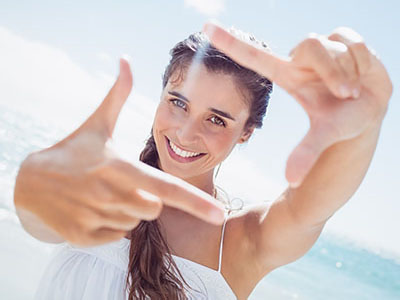The image features a woman taking a selfie with her hand held up against the sun, smiling at the camera while standing on a beach.