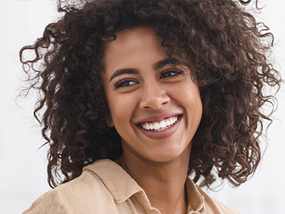 The image shows a person smiling at the camera with curly hair, wearing a beige top.